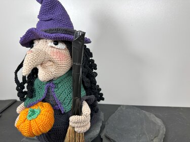 Gehäkelte Amigurumi‑Hexe mit lila Hut, grünem Umhang, schwarzem Haar, hält einen orangefarbenen Kürbis und einen Besen.