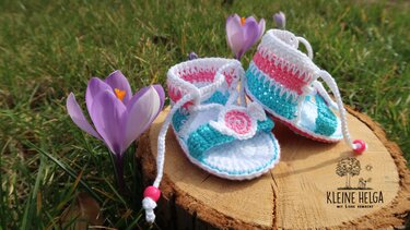 Gehäkelte Babysandalen in Pastellfarben mit Blüte auf Holzscheibe im Gras