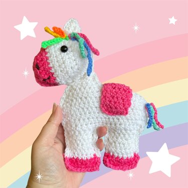 Weißes gehäkeltes Einhorn-Amigurumi mit Regenbogenmähne und pinken Akzenten, in einer Hand vor pastellfarbenem Regenbogenhintergrund.