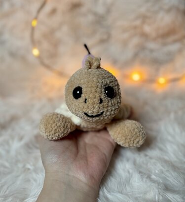 Beige gehäkelte Amigurumi-Schildkröte mit schwarzen Augen, in einer Hand gehalten vor unscharfem, beleuchtetem Hintergrund.