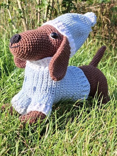 Gehäkelter Dackel-Amigurumi mit hellblauem Pullover und Mütze auf Gras
