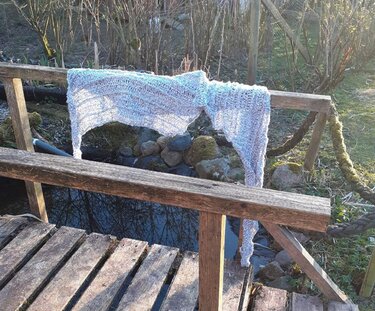 Hellgrauer gehäkelter Schal mit offenem Muster auf verwittertem Holzgeländer über einem Teich