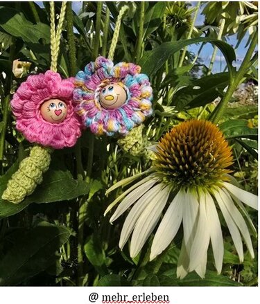 Nahaufnahme zweier gehäkelter Blumen-Anhänger mit bemalten Gesichtern, hängend an grünen Stielen