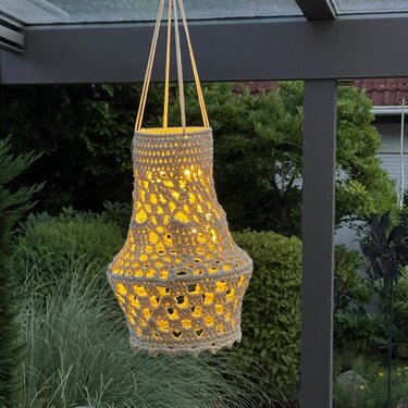 Gehäkelte hängende Laterne mit warmem Licht vor Gartenpflanzen