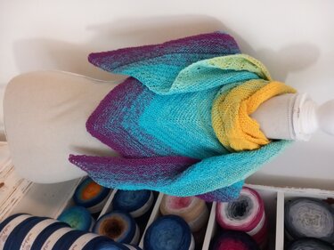 Gestricktes Dreieckstuch mit Farbverlauf in Lila, Türkis und Gelb auf einer Schneiderpuppe, Garnknäuel im Vordergrund.