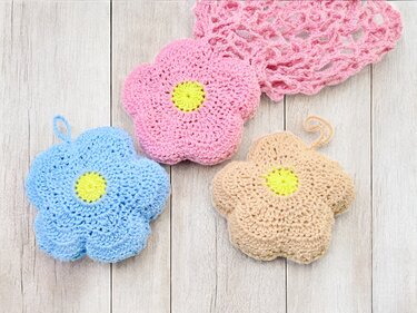 Three crocheted flower appliqués in pink, blue, and beige with yellow centers on a light wooden surface beside a pink openwork crochet fabric.