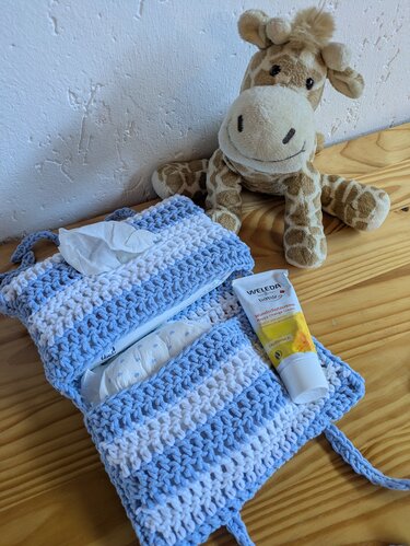 Gehäkelte blau-weiße Wickeltasche mit Windeln, Feuchttüchern und Pflegecreme; Stoffgiraffe im Hintergrund.