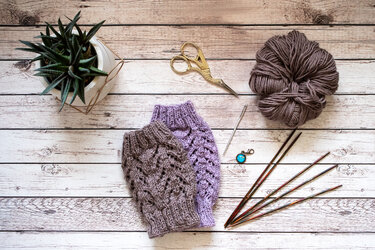 Zwei gestrickte Kinder-Beinstulpen, Wollknäuel, Stricknadeln und Schere auf hellem Holztisch.