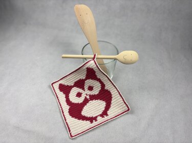 Quadratischer zweifarbiger Topflappen mit Eulen-Grafik und schmaler Borte, auf hellgrauem Hintergrund neben Küchenlöffeln