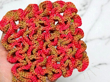 Nahaufnahme einer handgehäkelten Applikation in Rot-, Orange- und Goldtönen mit Wellenmuster, gehalten von einer Hand.