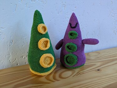 Zwei farbige, gehäkelte Kegel-Amigurumi mit applizierten Ringen und gesticktem Gesicht auf Holzuntergrund