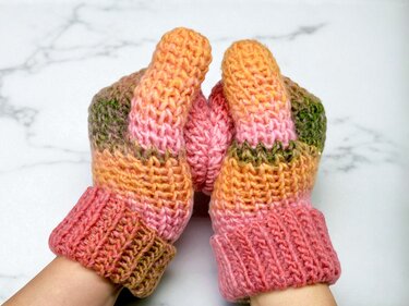 Hände in gehäkelten Fäustlingen mit weichem Farbverlauf aus Rosa, Orange und Grün.