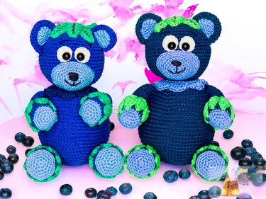 Crochet blueberry teddy bears in navy and light blue with green leaf edging posed with real blueberries