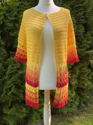 Gehäkelte gelbe Damenjacke mit Farbverlauf an Ärmeln und Saum auf einer Schneiderpuppe vor grüner Hecke.