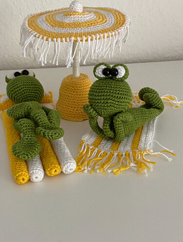 Zwei gehäkelte grüne Amigurumi-Frösche auf gelben und weißen Strandtüchern unter gestreiftem Sonnenschirm