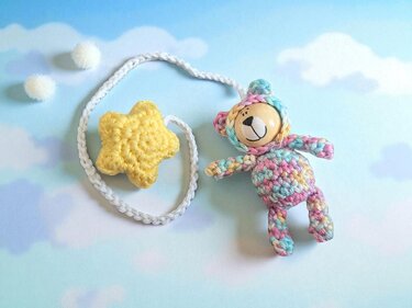 Gehäkelter Teddybär mit Holzkopf und pastellfarbenem Körper neben gelbem Häkelstern an weißer Kordel auf blauem Wolkenhintergrund