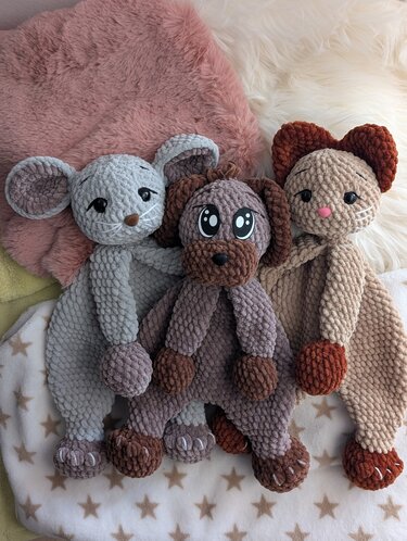 Drei gehäkelte Schmusetücher in Tierform: graue Maus, brauner Hund und beigefarbene Katze auf einer Decke mit Sternmuster