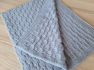 Gefaltete hellgraue gestrickte Babydecke mit Strukturfeldern und Zopfmuster auf hellem Holzhintergrund