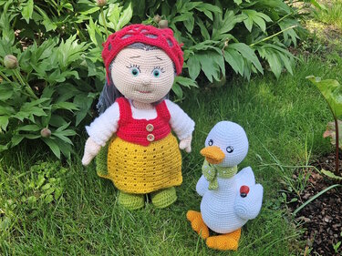 Gehäkelte Amigurumi-Puppe mit rotem Haarnetz, roter Weste und gelbem Rock neben einer weißen Ente mit grünem Schal auf Rasen