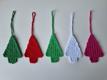 Fünf gestrickte Baum-Anhänger (rot, zwei Grüntöne, weiß, pink) mit Schlaufen auf hellem Hintergrund