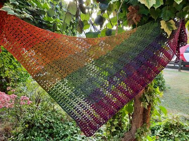 Häkel-Dreieckstuch mit Farbverlauf von Orange über Grün zu Violett, luftiges Spitzenmuster, an Zweigen aufgehängt