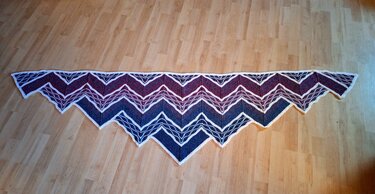 Gestricktes Dreieckstuch mit Zickzackmuster und Farbverlauf von Dunkelblau zu Weinrot, mit weißen Konturlinien auf Holzboden.