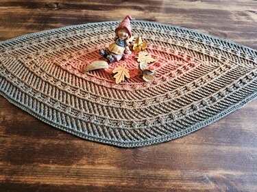 Gehäkelter ovaler Tischläufer mit warmem orangefarbenem Zentrum und grauem Rand, dekoriert mit kleiner Wichtelfigur und Holzblättern auf Holztisch