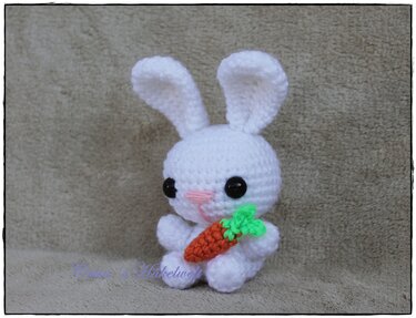 Weißer gehäkelter Amigurumi-Hase mit schwarzen Augen, rosafarbener Nase und einer kleinen orange-grünen Karotte.