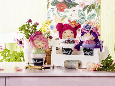 Drei Nadelkissen als Stoffpuppen in bemalten Blumentöpfen auf einem Tisch mit Blumen und Nähgarn