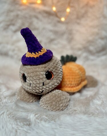 Samtige gehäkelte Amigurumi-Schildkröte mit lila Hexenhut und orangefarbener Schale auf hellem Plüschuntergrund