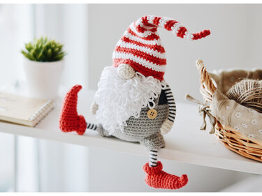 Gehäkelter Wichtel mit rot-weiß gestreifter Mütze, weißem Bart und roten Schuhen auf einem Regal