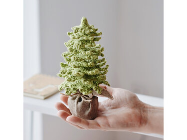 Handgehäkelter Mini-Tannenbaum in hellem Grün im Jutesäckchen, in einer Hand gehalten.
