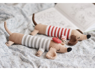 Zwei gehäkelte Dackel-Amigurumi in gestreiften Pullovern, einer mit roter Baskenmütze, auf geblümter Decke.