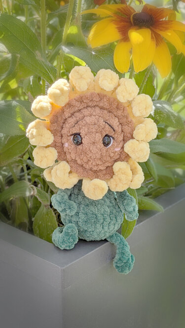 Gehäkeltes Sonnenblumen-Amigurumi mit braunem Kopf und gelben Blütenblättern auf grauem Pflanzkübel