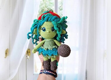 Gehäkelte grüne Amigurumi‑Puppe mit blaugrünem Lockenhaar und kleinem braunen Ball, gehalten in einer Hand.