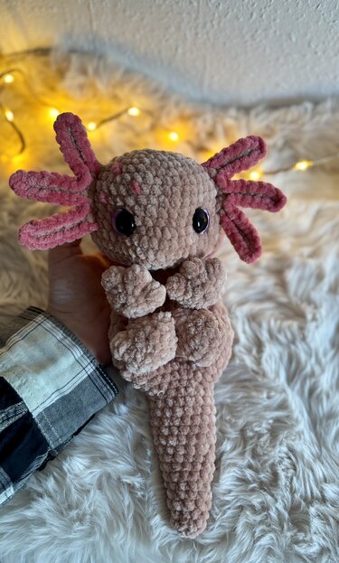 Hand hält roséfarbenes gehäkeltes Axolotl‑Amigurumi mit rosa Kiemen vor flauschigem Untergrund und Lichterkette