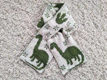 Child-sized knitted scarf with green dinosaur motifs and small star repeats on a cream background
