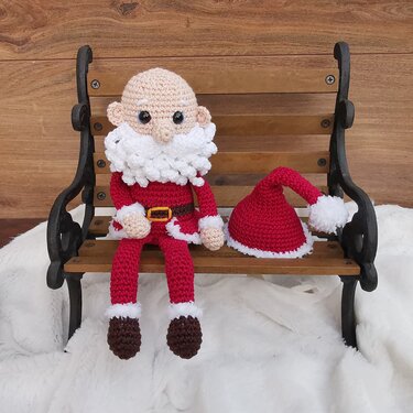 Gehäkelte Amigurumi-Weihnachtsmann-Puppe mit weißem Bart und rotem Anzug auf einer Holzbank, die rote Zipfelmütze liegt daneben