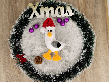 Gehäkelte weiße Vogel-Amigurumi mit roter Mütze und gelben Füßen in grün-weißem Kranz neben violetten Christbaumkugeln