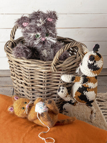 Drei gehäkelte Katzen-Amigurumi: graue Katze im Weidenkorb, orange Katze auf Kissen mit Wollknäuel, gefleckte Katze daneben.
