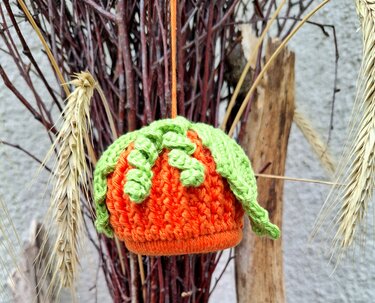 Kleiner gehäkelter oranger Kürbis mit grünen Blättern, aufgehängt vor Zweigen und Weizenähren