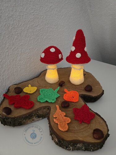 Zwei gehäkelte rot-weiße Fliegenpilz-Lampen auf Holzscheibe, umgeben von gehäkelten Herbstblättern und Kastanien