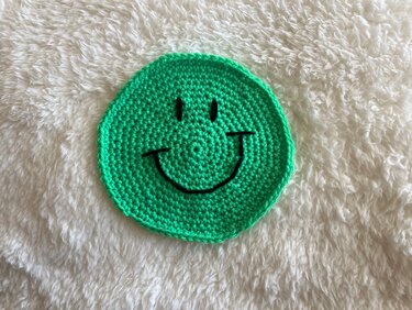 Runder grüner, gehäkelter Smiley-Untersetzer auf weißem flauschigem Stoff.