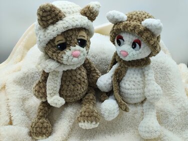 Zwei gehäkelte Amigurumi-Kätzchen in Braun und Creme mit Mützen und Schals auf flauschiger Decke.