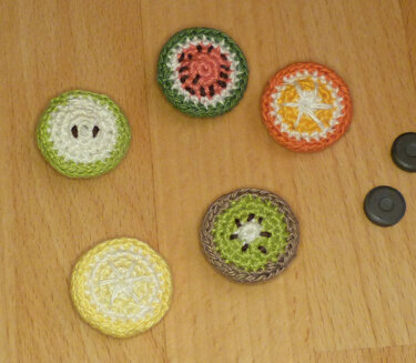 Fünf gehäkelte runde Fruchtscheiben‑Magnete (Wassermelone, Orange, Kiwi, Zitrone, Apfel) auf Holzuntergrund.