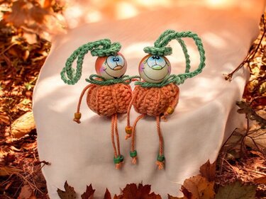 Zwei gehäkelte orangefarbene Kürbis-Anhänger mit bemalten Perlenköpfen und grünen Aufhängeschlaufen auf hellem Stoff vor Herbstlaub