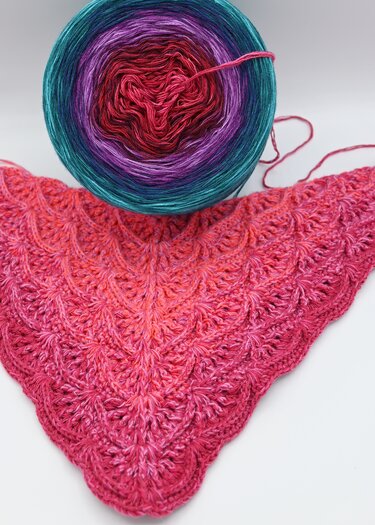 Nahaufnahme eines pink-rot gefärbten, gehäkelten Dreieckstuchs mit Muschelstruktur; darüber ein Farbverlaufsgarnknäuel in Türkis, Lila und Rot auf hellem Hintergrund.