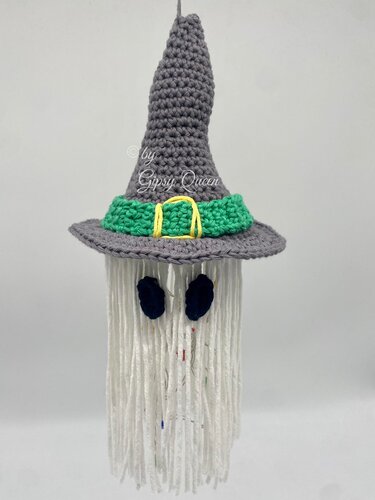 Gehäkelter Geist mit spitzem grauen Hexenhut, grünem Band, gelber Schnalle und weißen Garnfransen vor hellem Hintergrund.