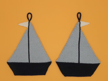 Zwei gestrickte Segelboot-Topflappen mit hellgrauen Segeln, dunklen Rümpfen, weißen Wimpeln und Aufhängeschlaufen auf orangefarbenem Hintergrund.