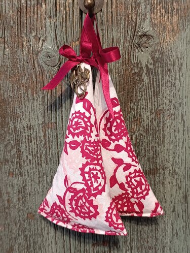 Zwei weiße Stoffkegel mit rotem Blumenmuster, zusammengebunden mit rotem Satinband und kleinem Drachenanhänger, hängend an einem hölzernen Haken.